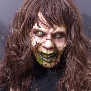 May include: A close-up of a horror movie mask with long brown hair. The mask has green and white makeup, and the eyes are painted with green and yellow accents.