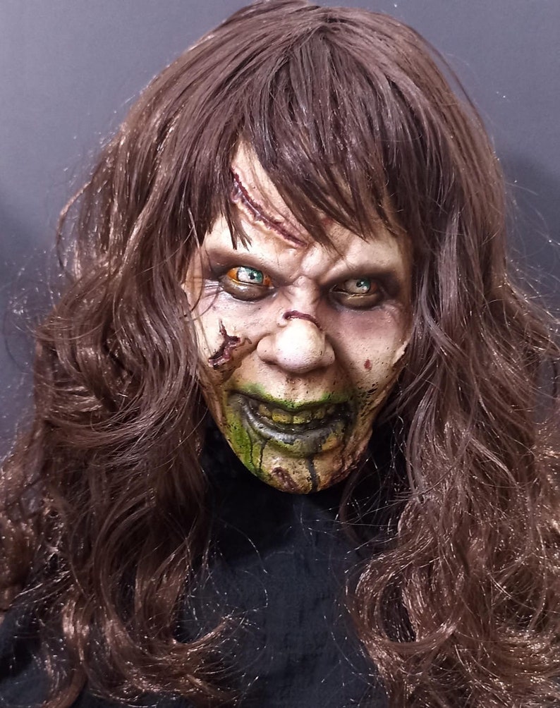 May include: A close-up of a horror movie mask with long brown hair. The mask has green and white makeup, and the eyes are painted with green and yellow accents.