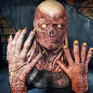 May include: A detailed, reddish-brown monster mask with glowing yellow eyes and yellow-tipped fingernails. The mask has a textured, veiny appearance, and the hands are raised in front of the face. The overall theme is horror and Halloween.