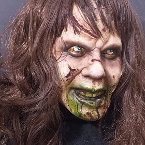 May include: A close-up of a horror movie mask with long brown hair. The mask has a pale green and white face with green dripping paint, a wide smile, and large eyes with green and orange irises.