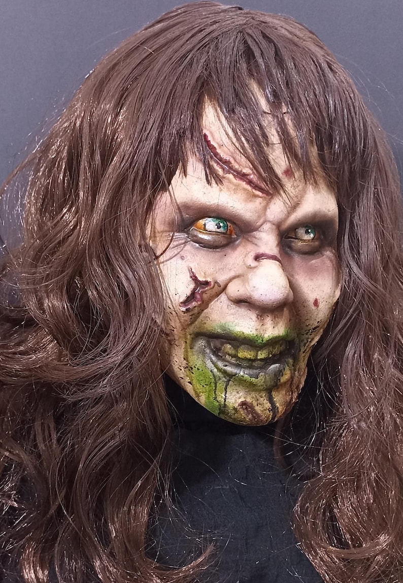 May include: A close-up of a horror movie mask with long brown hair. The mask has a pale green and white face with green dripping paint, a wide smile, and large eyes with green and orange irises.