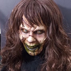 May include: A close-up of a horror movie mask with long brown hair. The mask is made of latex and has a realistic, grotesque appearance. The mask has green and black paint around the mouth and eyes, and the eyes are painted a bright green color.