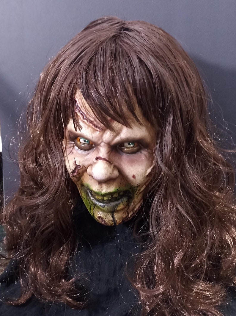 May include: A close-up of a horror movie mask with long brown hair. The mask is made of latex and has a realistic, grotesque appearance. The mask has green and black paint around the mouth and eyes, and the eyes are painted a bright green color.