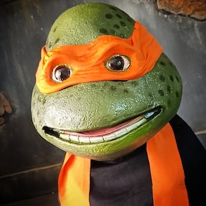 May include: A close-up of a green turtle mask with an orange eye mask and scarf. The mask has large, expressive eyes and a smiling mouth. The mask is worn over a black shirt.