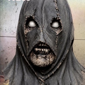 Könnte beinhalten: Eine verwitterte, dunkelgraue Maske mit einer genähten Kapuze und verfallenden Gesichtszügen. Die Maske hat weiße Augen und einen klaffenden Mund, der Zähne zeigt. Das Wort "NEW" ist oben in weißen Buchstaben zu sehen. Ein Halloween-Kostüm.