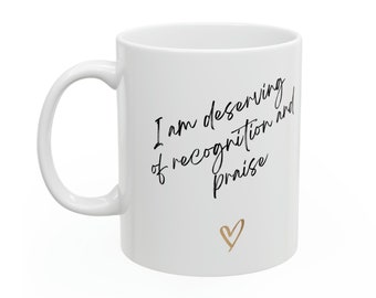 May include: White ceramic mug with a curved handle. The mug features the handwritten text "I am deserving of recognition and praise" in black script, along with a small gold heart. A simple and elegant design.