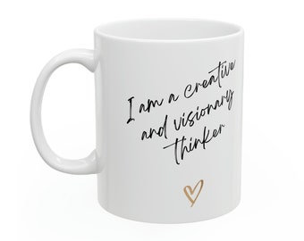May include: White ceramic mug with a curved handle. The mug features the text "I am a creative and visionary thinker" in black script, with a gold heart below the text. The mug is designed for hot beverages.