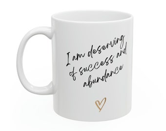May include: White ceramic mug with black text that reads "I am deserving of success and abundance" with a gold heart at the bottom.