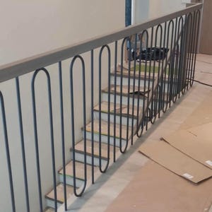 May include: A gray metal railing with a repeating arched design. The railing is installed along a staircase with light-colored steps. Cardboard sheets are on the floor near the stairs.