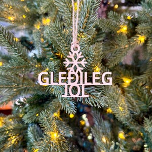 Puede incluir: Adorno navideño de madera con las palabras "GLEÐILEG JÓL" recortadas. El adorno tiene forma de copo de nieve y está colgado de una cuerda. El adorno cuelga de un árbol de Navidad con luces.