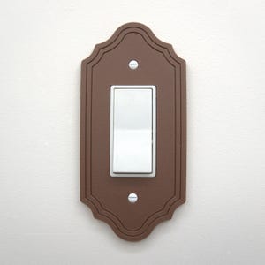 May include: A brown, decorative light switch cover with a white rocker switch. The cover has a scalloped edge and is secured with two silver screws. The switch is in the on position.