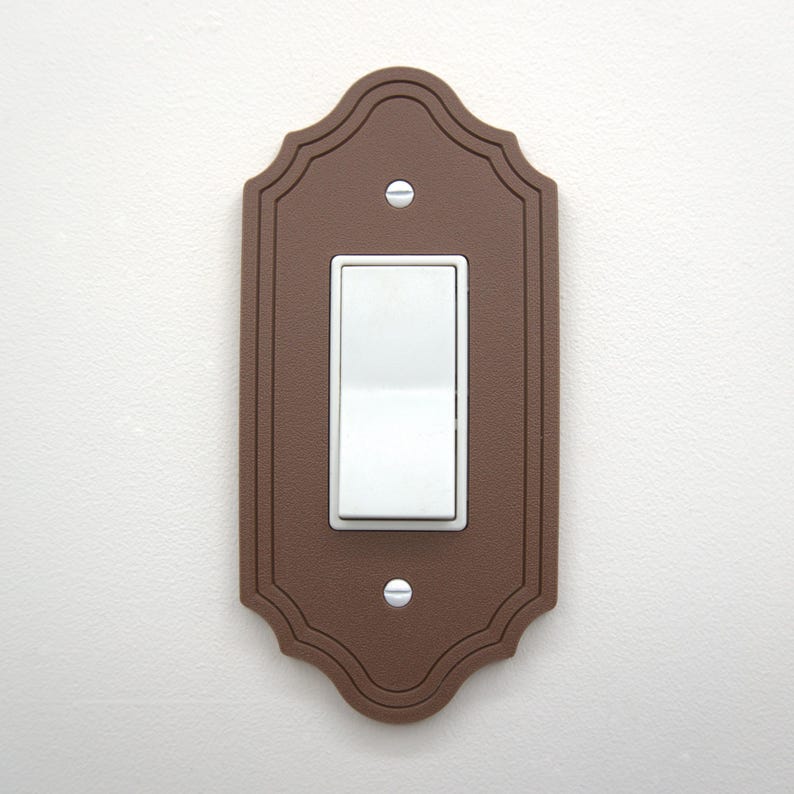 May include: A brown, decorative light switch cover with a white rocker switch. The cover has a scalloped edge and is secured with two silver screws. The switch is in the on position.