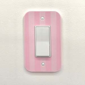 Op de afbeelding: Een lichtroze en wit gestreepte lichtschakelaar cover met een witte tuimelschakelaar. De cover is rechthoekig met afgeronde hoeken en is gemonteerd op een witte muur. Twee zilveren schroeven bevestigen de cover.