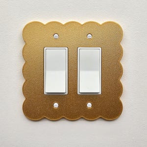 May include: A gold-coloured, scalloped-edge light switch plate with two white light switches. The plate is mounted on a white wall and secured with silver screws. The plate has a textured surface.