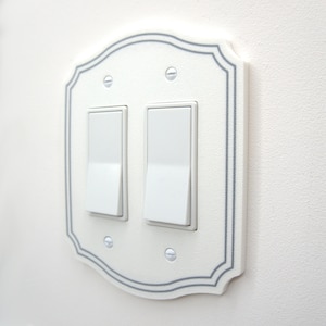 May include: A white double light switch wall plate with a decorative scalloped edge and a thin gray border. The rectangular plate features two white rocker switches and is secured to the wall with four small, silver-colored metal screws.