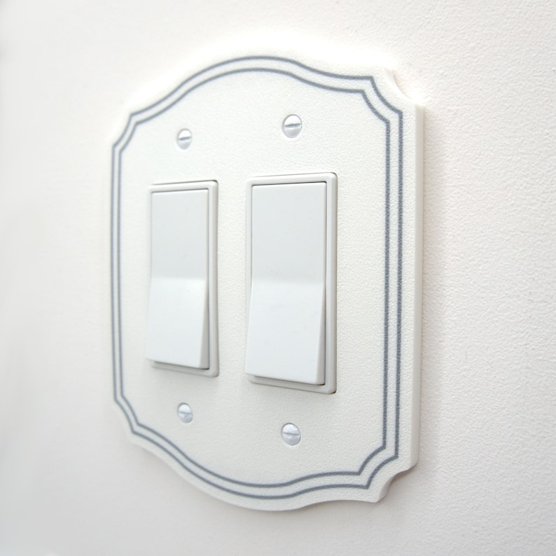 May include: A white double light switch wall plate with a decorative scalloped edge and a thin gray border. The rectangular plate features two white rocker switches and is secured to the wall with four small, silver-colored metal screws.
