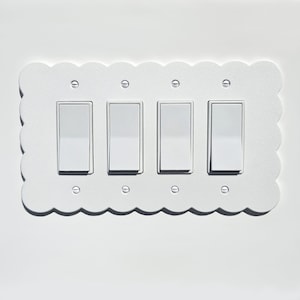 May include: A white, scalloped-edge light switch plate with four rectangular light switches. The plate is mounted on a white wall. The switches are in the on position.