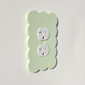 May include: A light green wall plate with two standard electrical outlets. The plate has a scalloped edge and is made of a textured material.