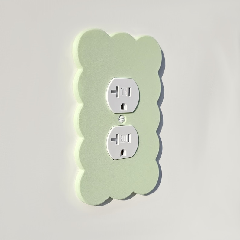 May include: A light green wall plate with two standard electrical outlets. The plate has a scalloped edge and is made of a textured material.