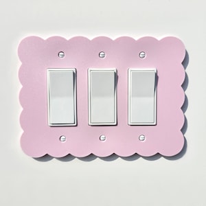 May include: A pink light switch plate with three white toggle switches. The plate has a scalloped edge and is designed to look like a cookie.