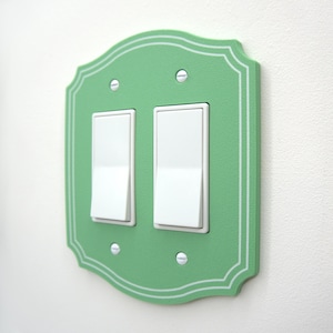 May include: A mint green double light switch wall plate with a scalloped edge and white pinstripe detailing. The rectangular cover features two white rocker switches and is secured to the wall with four silver-colored metal screws.