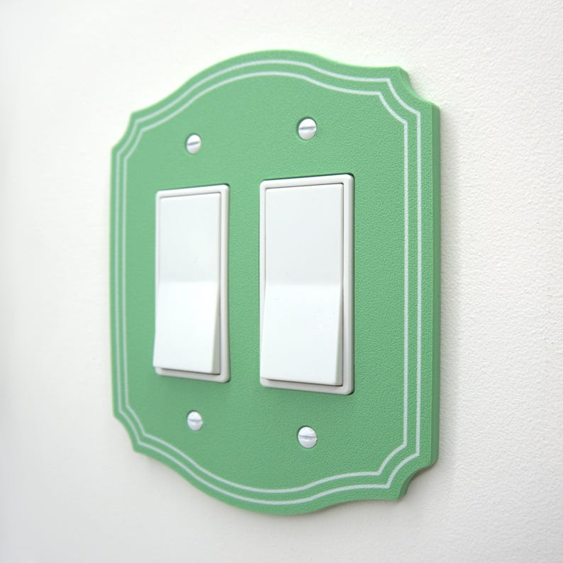 May include: A mint green double light switch wall plate with a scalloped edge and white pinstripe detailing. The rectangular cover features two white rocker switches and is secured to the wall with four silver-colored metal screws.