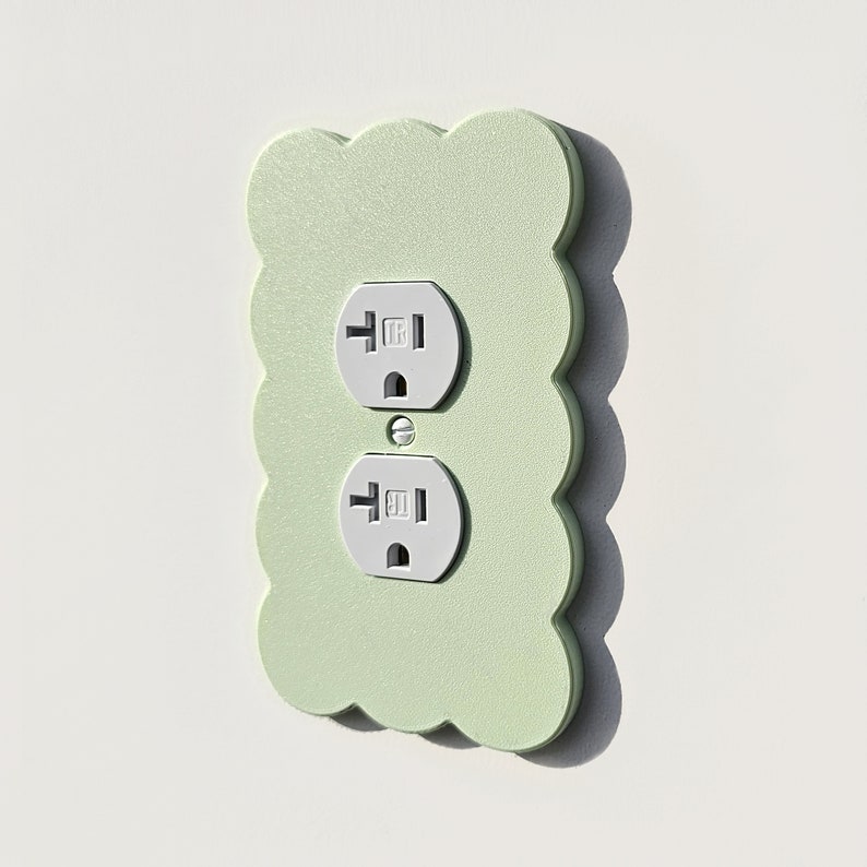 May include: A light green outlet cover with a scalloped edge and two white electrical outlets.