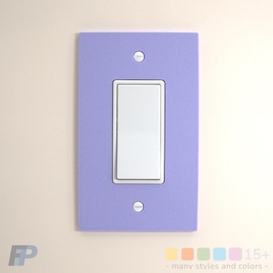 May include: A rectangular light switch cover plate in a solid matte lavender finish with a textured surface. The plate features a central vertical opening for a white rocker switch and is secured by two silver-toned metal screws at the top and bottom.