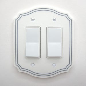 May include: A white double rocker light switch wall plate with a scalloped edge and a thin gray border. The plate features two white rectangular rocker switches and is secured to the wall with four silver-colored metal screws at the corners.