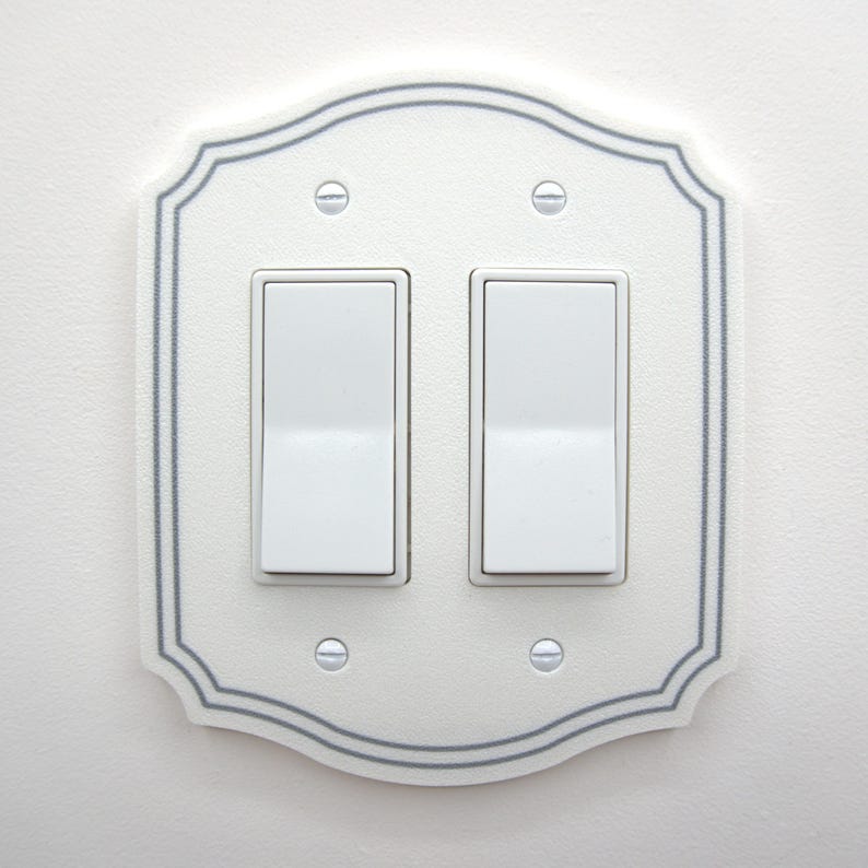 May include: A white double rocker light switch wall plate with a scalloped edge and a thin gray border. The plate features two white rectangular rocker switches and is secured to the wall with four silver-colored metal screws at the corners.