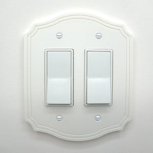 May include: A white, decorative light switch plate with two toggle switches. The plate has a scalloped edge and is mounted on a white wall. Four screws secure the plate. The switches are in the off position.