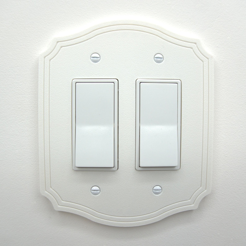 May include: A white, decorative light switch plate with two toggle switches. The plate has a scalloped edge and is mounted on a white wall. Four screws secure the plate. The switches are in the off position.