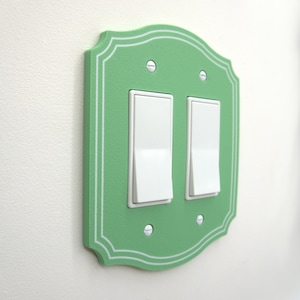 May include: A light green double light switch wall plate with a decorative scalloped edge and white pinstripe detailing. The plate features two white rectangular rocker switches and is secured to the wall with four visible silver metal screws.
