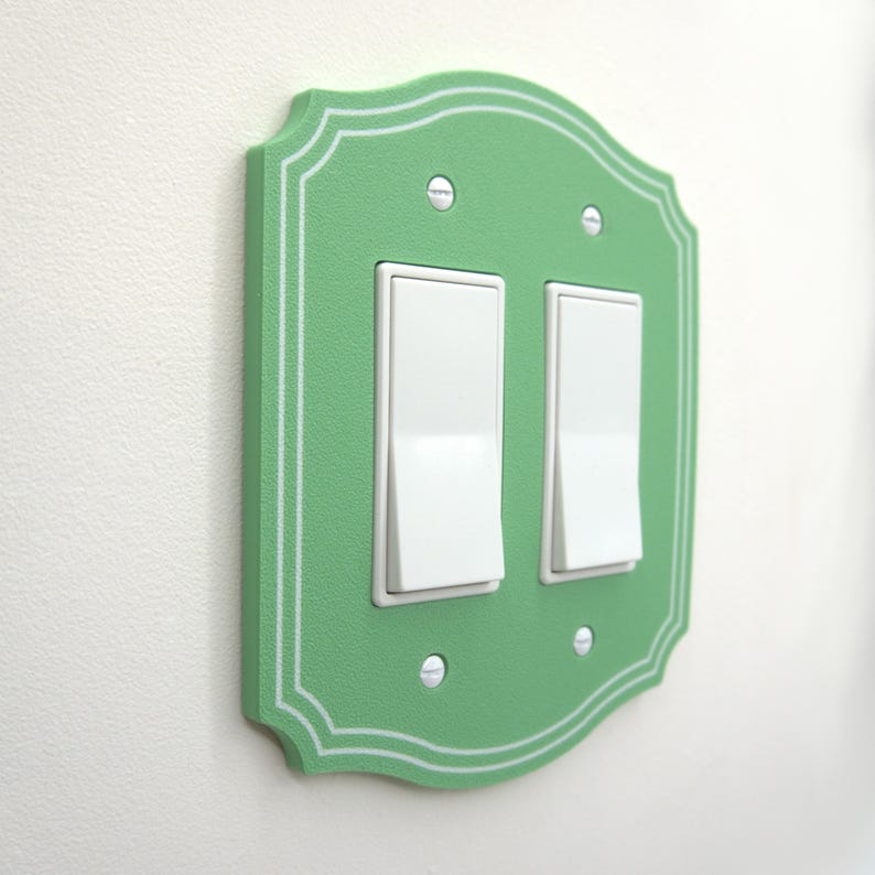 May include: A light green double light switch wall plate with a decorative scalloped edge and white pinstripe detailing. The plate features two white rectangular rocker switches and is secured to the wall with four visible silver metal screws.
