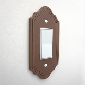 May include: A brown, decorative light switch cover with a white toggle switch. The cover has a scalloped edge and is mounted on a white wall. The switch is in the on position.