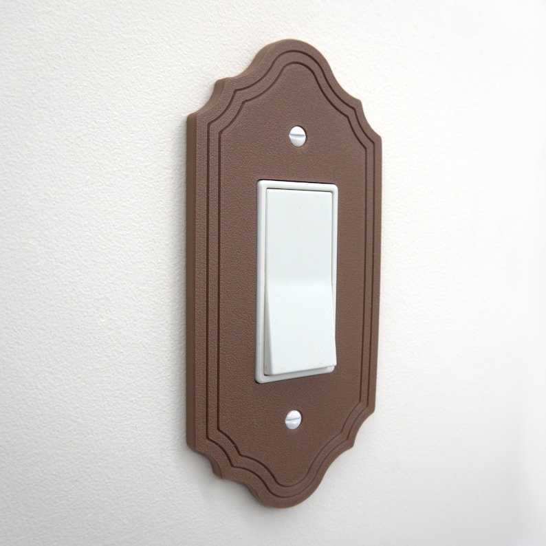May include: A brown, decorative light switch cover with a white toggle switch. The cover has a scalloped edge and is mounted on a white wall. The switch is in the on position.