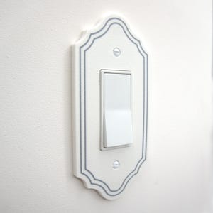 May include: A white light switch cover plate with a decorative scalloped edge and a thin gray border line. The rectangular plate features a central rocker switch and is secured to the wall with two visible silver-toned metal screws.
