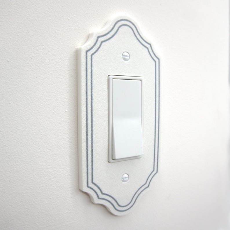 May include: A white light switch cover plate with a decorative scalloped edge and a thin gray border line. The rectangular plate features a central rocker switch and is secured to the wall with two visible silver-toned metal screws.