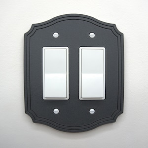 May include: A decorative black double light switch cover with a scalloped edge. The cover has two white rocker light switches and is secured with four silver screws. The cover is mounted on a white wall.