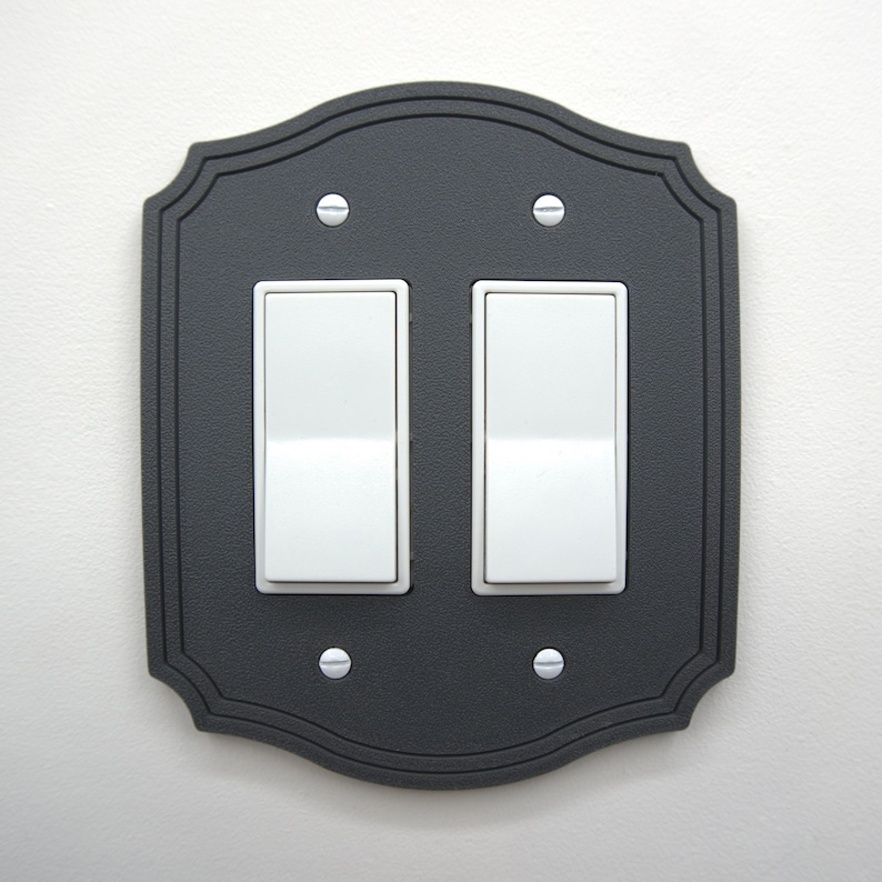 May include: A decorative black double light switch cover with a scalloped edge. The cover has two white rocker light switches and is secured with four silver screws. The cover is mounted on a white wall.
