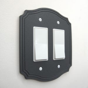 May include: A decorative, dark gray light switch cover with two white rocker switches. The cover has a scalloped edge and is secured with four silver screws. The switches are in the on position.