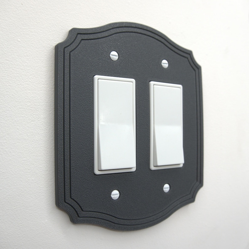 May include: A decorative, dark gray light switch cover with two white rocker switches. The cover has a scalloped edge and is secured with four silver screws. The switches are in the on position.