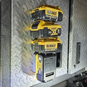 Puede incluir: Cuatro baterías DeWalt 20V MAX de iones de litio de color amarillo y negro están apiladas una encima de la otra. Las baterías están sujetas a una pared con una tira magnética.