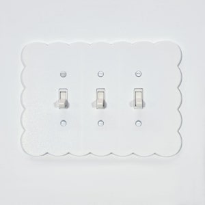 Könnte beinhalten: Weiße Lichtschalterplatte mit drei Kippschaltern. Die Platte hat einen gewellten Rand und ist an einer weißen Wand montiert.