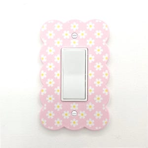 Puede incluir: Una placa de interruptor de luz rosa con un patrón de margaritas. La cubierta tiene un borde festoneado y un interruptor basculante blanco en el centro. Las margaritas son blancas con centros amarillos.