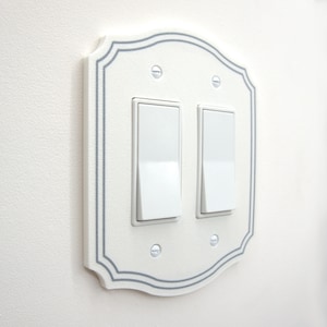 May include: A white double rocker light switch wall plate with a scalloped edge and a thin gray border. The plate features two white rectangular rocker switches and is secured to the wall with four small, silver-colored metal screws.