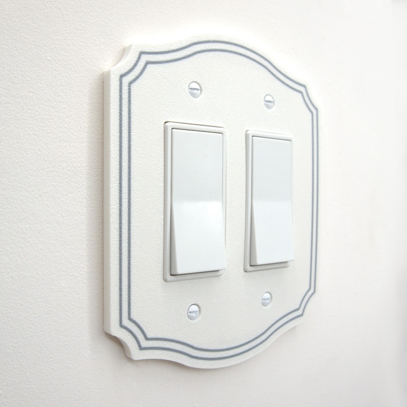 May include: A white double rocker light switch wall plate with a scalloped edge and a thin gray border. The plate features two white rectangular rocker switches and is secured to the wall with four small, silver-colored metal screws.