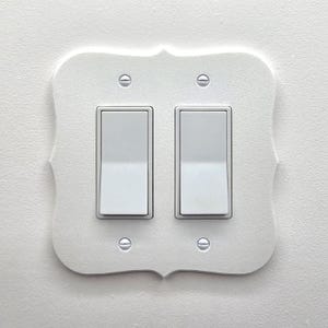以下が含まれることがあります： 2つのトグルスイッチが付いた白い装飾的なライトスイッチカバー。カバーはスカラップエッジで、白い壁に取り付けられています。スイッチとネジはシルバーです。