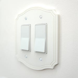 May include: A white, decorative double light switch cover with a scalloped edge design. The cover features two white rocker light switches and is mounted on a white wall. The screws are visible.