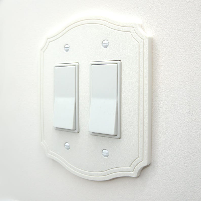 May include: A white, decorative double light switch cover with a scalloped edge design. The cover features two white rocker light switches and is mounted on a white wall. The screws are visible.